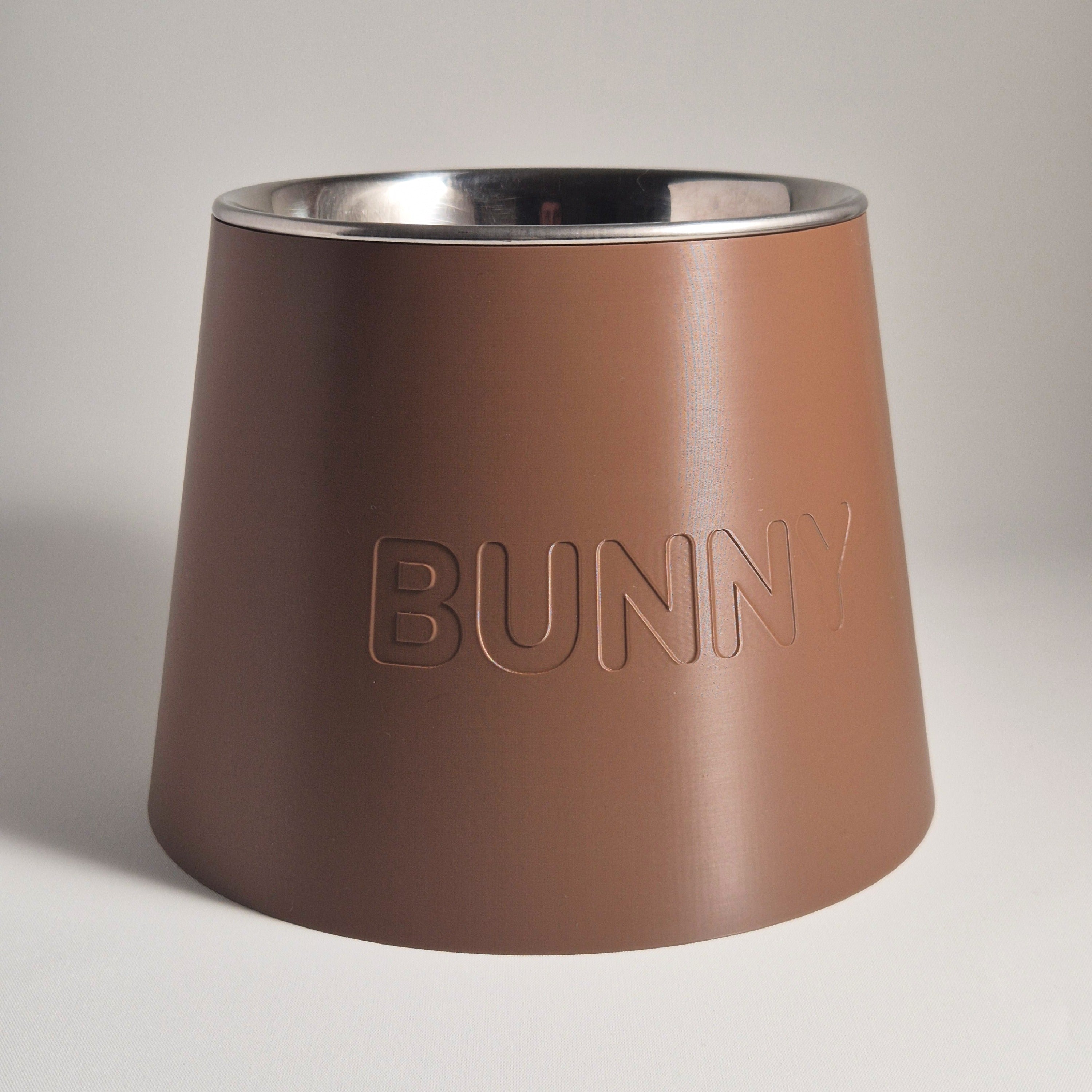 A conical pet feeder or bowl with a matte brown exterior and a polished stainless steel interior, featuring the word 'BUNNY' debossed on the front, set against a neutral light gray background.