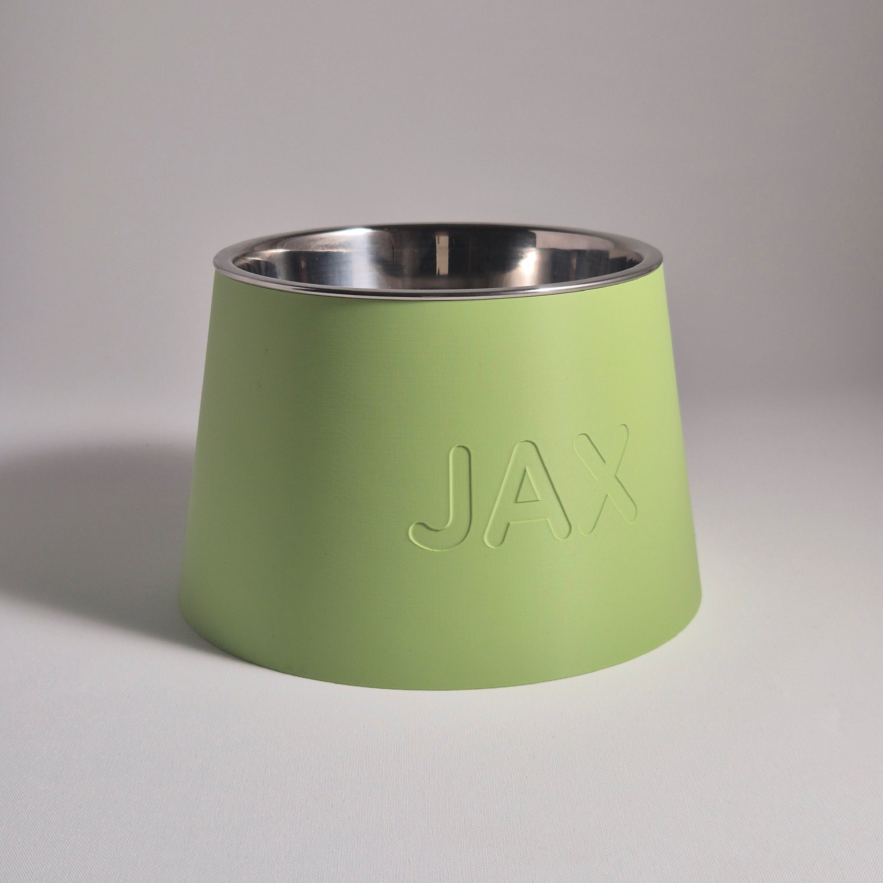 A light green pet bowl with a stainless steel interior and the name 'JAX' debossed on the front, set against a neutral gray background.