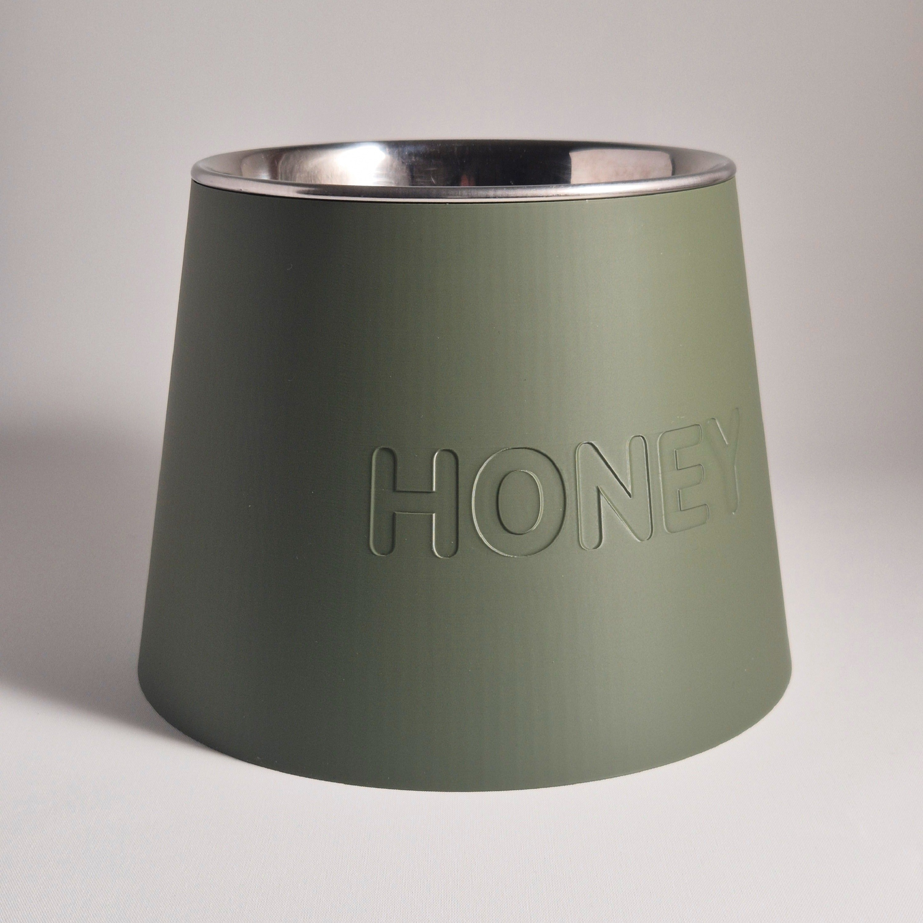 An olive green pet feeder bowl with a brushed silver stainless steel interior, featuring the word 'HONEY' debossed on the front, set against a neutral light gray background.