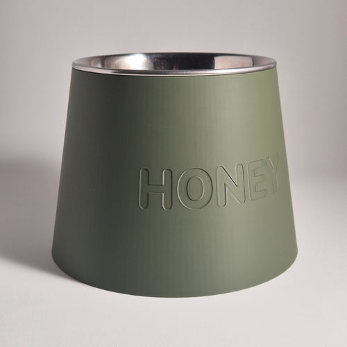 An olive green pet feeder bowl with a brushed silver stainless steel interior, featuring the word 'HONEY' debossed on the front, set against a neutral light gray background.