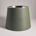 An olive green conical pet bowl with a brushed silver stainless steel interior, featuring the word 'HONEY' debossed on the front, set against a neutral gray background.