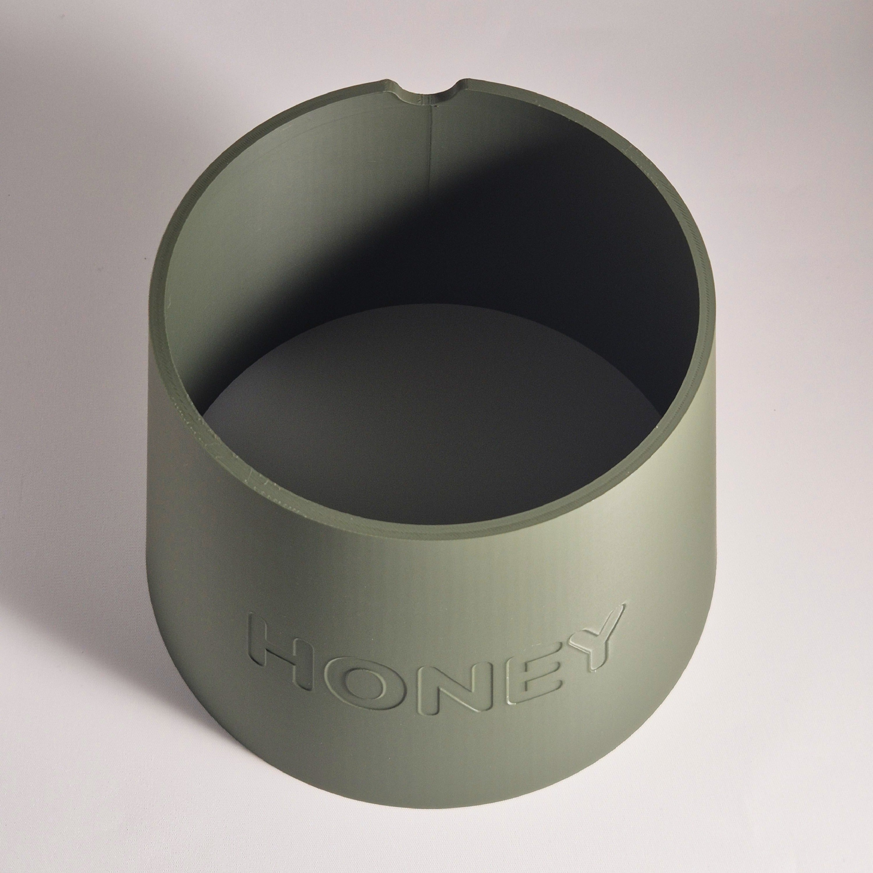 An olive green cylindrical container with a small notch at the top rim, featuring the word 'HONEY' debossed on the front, set against a neutral light gray background.